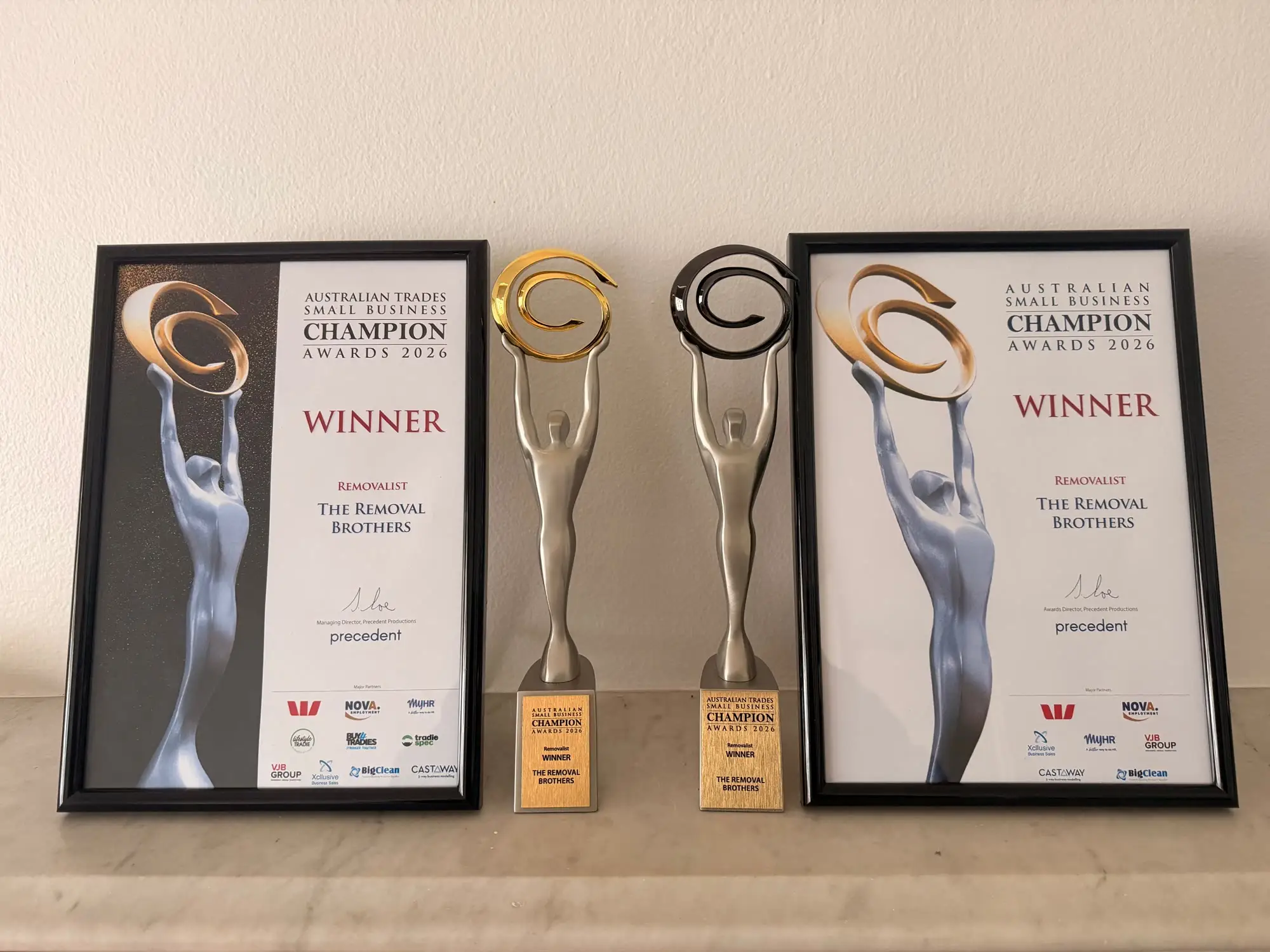 Both 2026 Removalist winner certificates framed beside their two Champion Awards trophies