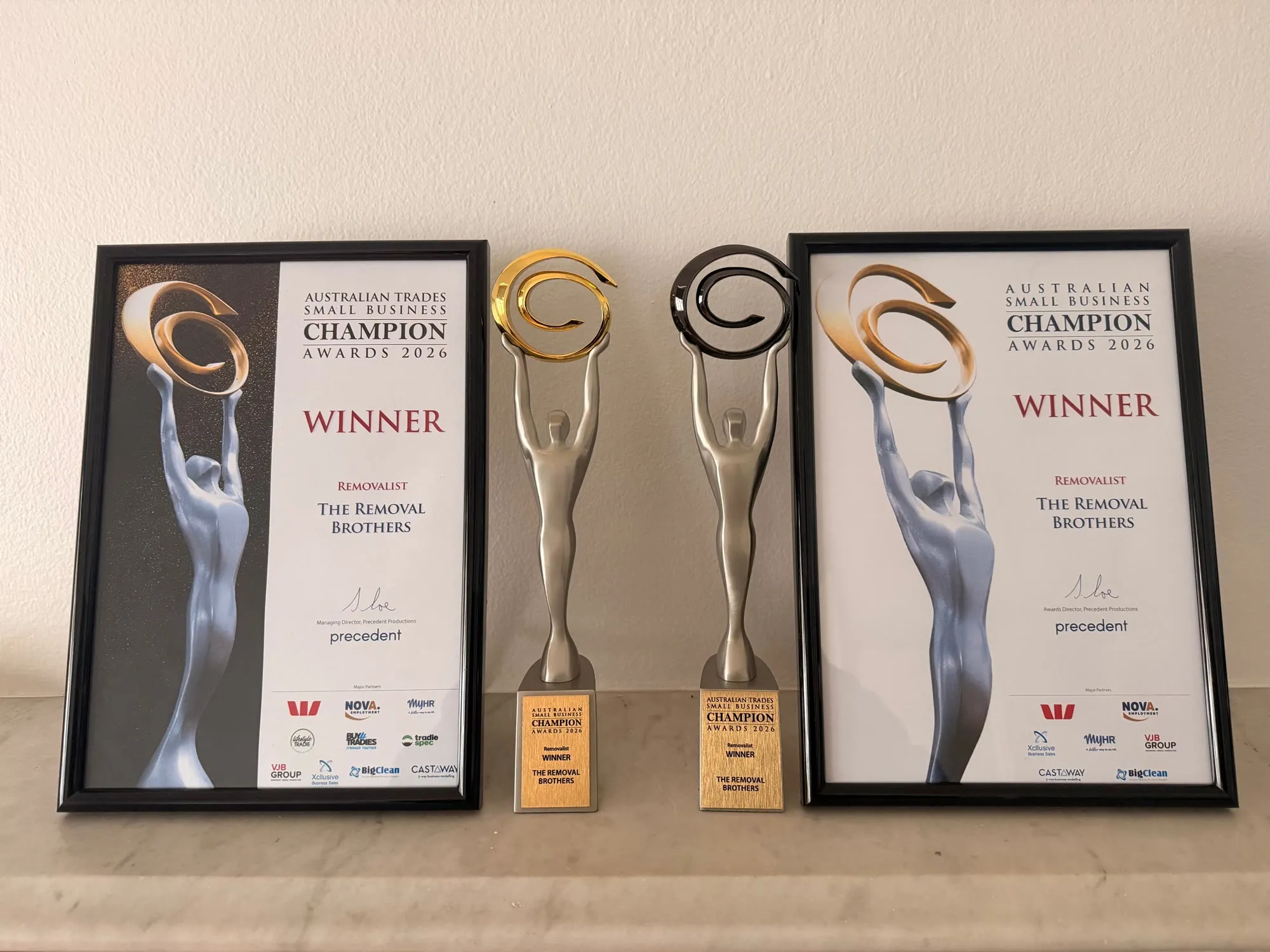 Both 2026 Removalist winner certificates framed beside their two Champion Awards trophies