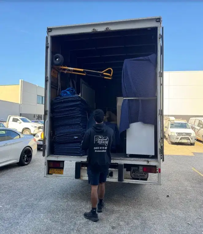 A Removal Brothers crew member loading a truck stacked with folded moving blankets, a trolley and wrapped furniture