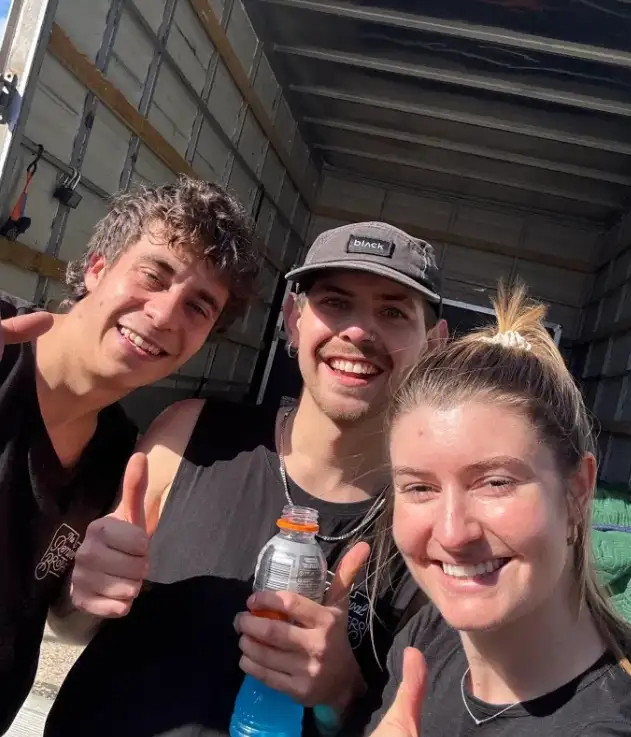 Three people photographed together in front of an open removal truck on a sunny day