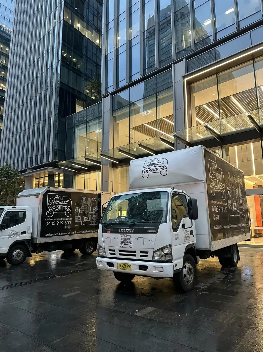 Two branded Removal Brothers trucks parked outside a Sydney office tower on a wet forecourt